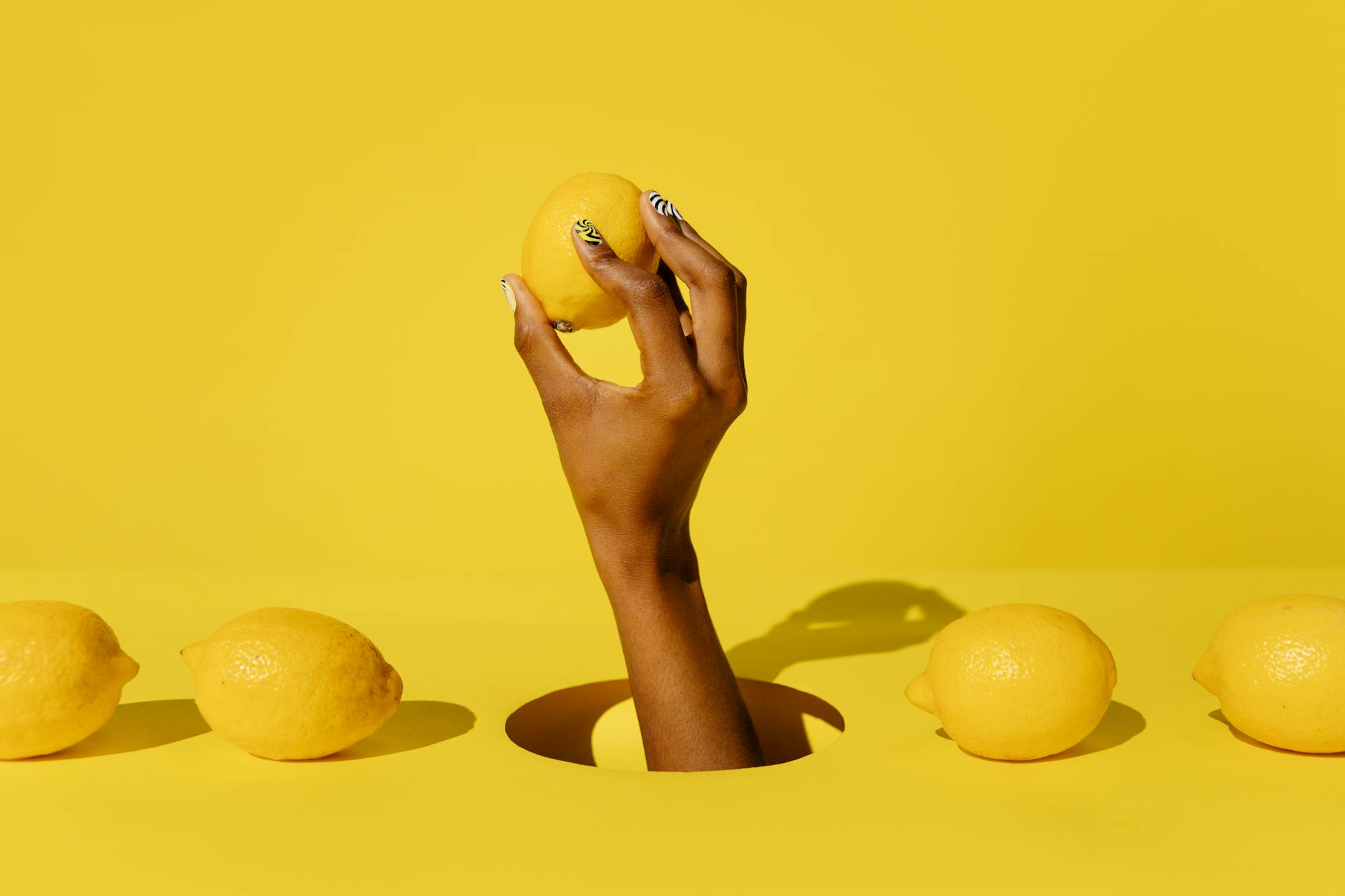 Hand holding a fresh lemon on a soft pink background surrounded by additional lemons