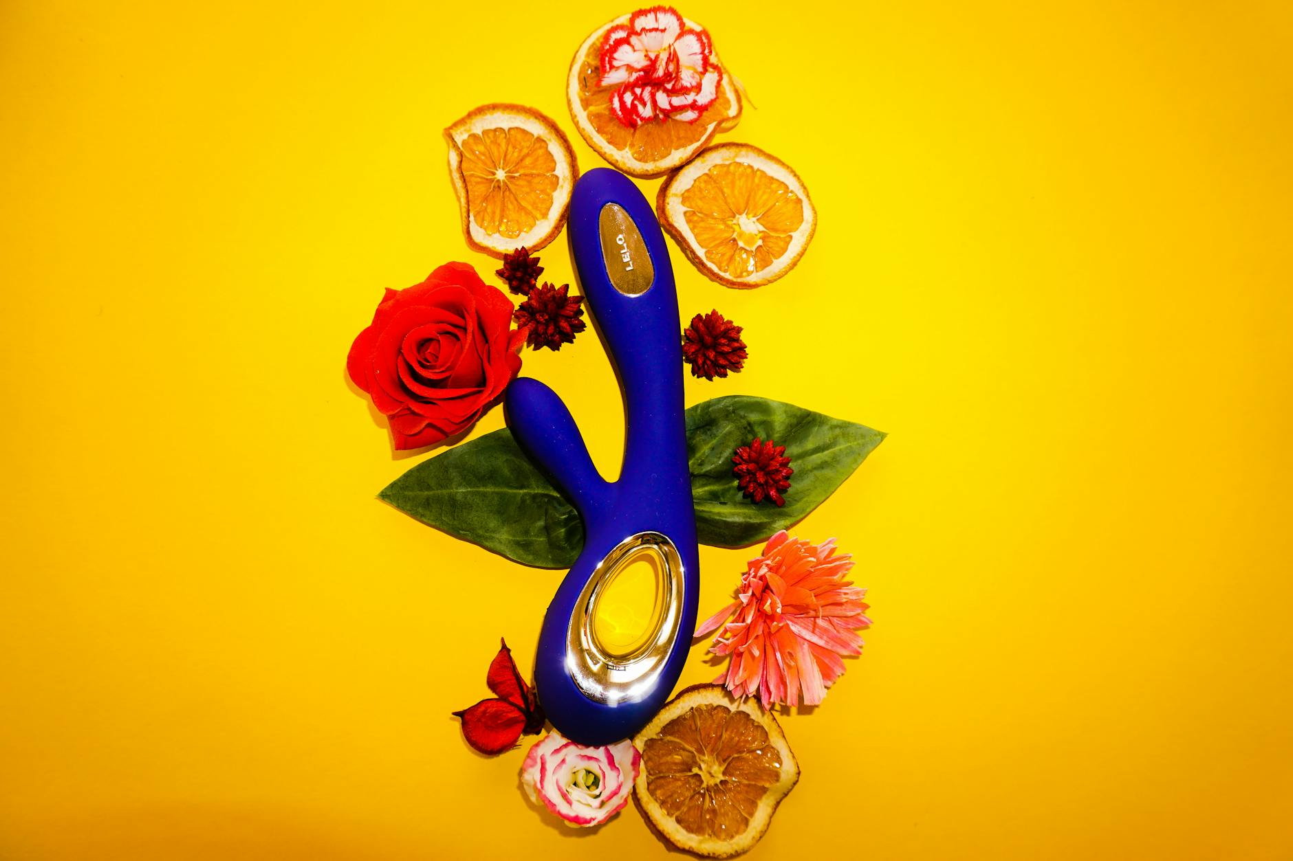A blue clitoral vibrator surrounded by flowers and citrus fruit on a bright yellow background